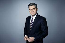 Jim Acosta wearing black coat and tie, and in a plain gradient grey background