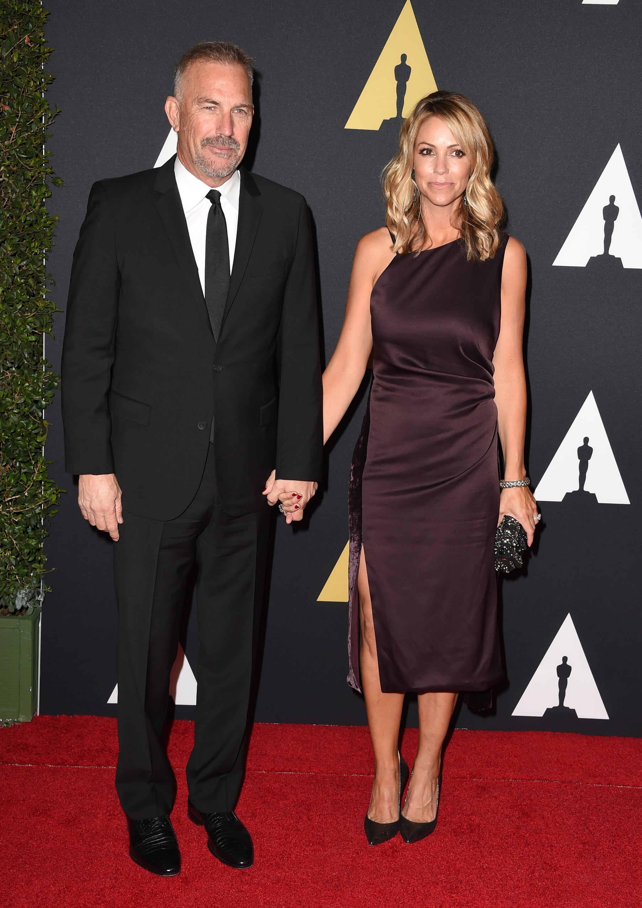 Christine Baumgartner wearing a black silk dress, holding hands with Kevin Cosner at an award show