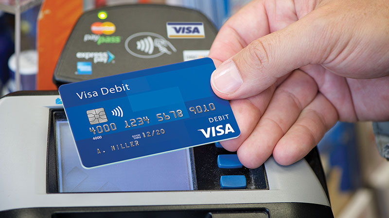 A blue Visa debit card held by hand near a swipe machine