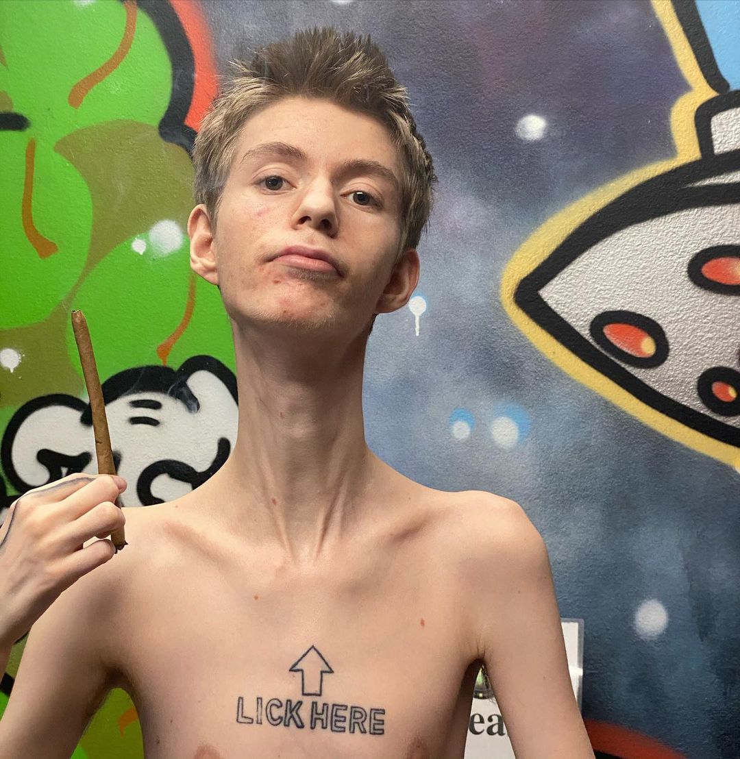Daddy Long Neck with chest tattoo of words ‘lick here’ in uppercase and above it a thick arrow pointing upward