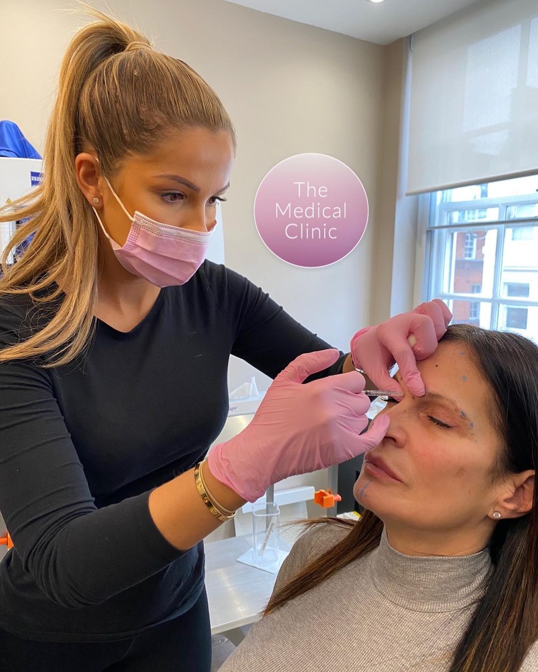 Woman in ponytail wearing pink facemask and pink surgical gloves injects Botox to a woman at The Medical Clinic in London