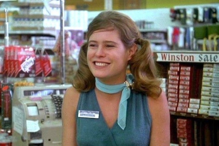 Sarah Holcomb role as a cashier wearing a blue-green dress and a blue scarf on her neck