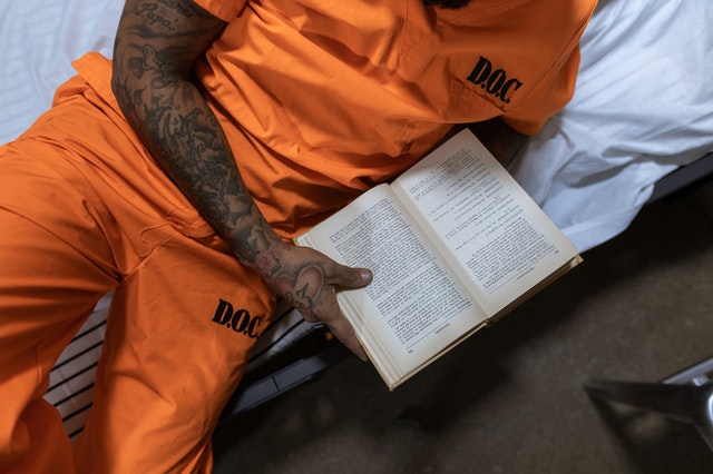 Tattooed male prisoner in D.O.C. orange uniform reading a book inside his cell