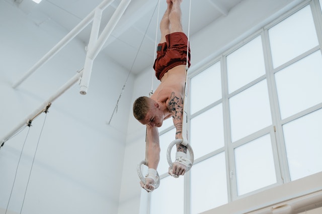 Shirtless male gymnast in red gym shorts with tattoos on left arm doing upside down ring pull-ups