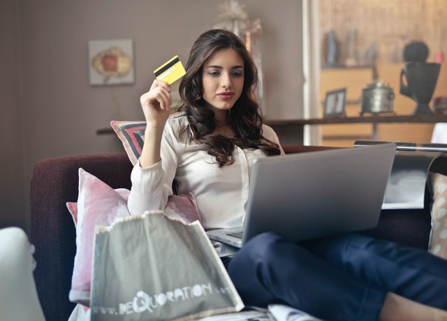 Woman holds a yellow credit card with right hand while doing online shopping from her couch on her silver laptop