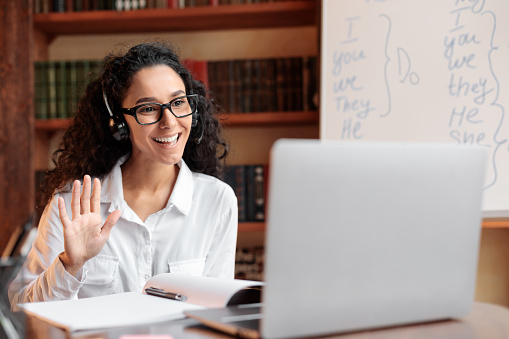 A woman wearing eyeglasses and headphones, signaling a greeting in front of her laptop during an online class