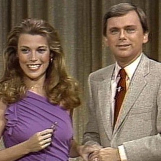 Vanna White in violet sleeveless gown and Pat Sajak in gray suit and red tie