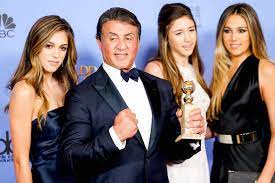 Sylvester Stallone holding a trophy with his three daughters, and a blue background behind them