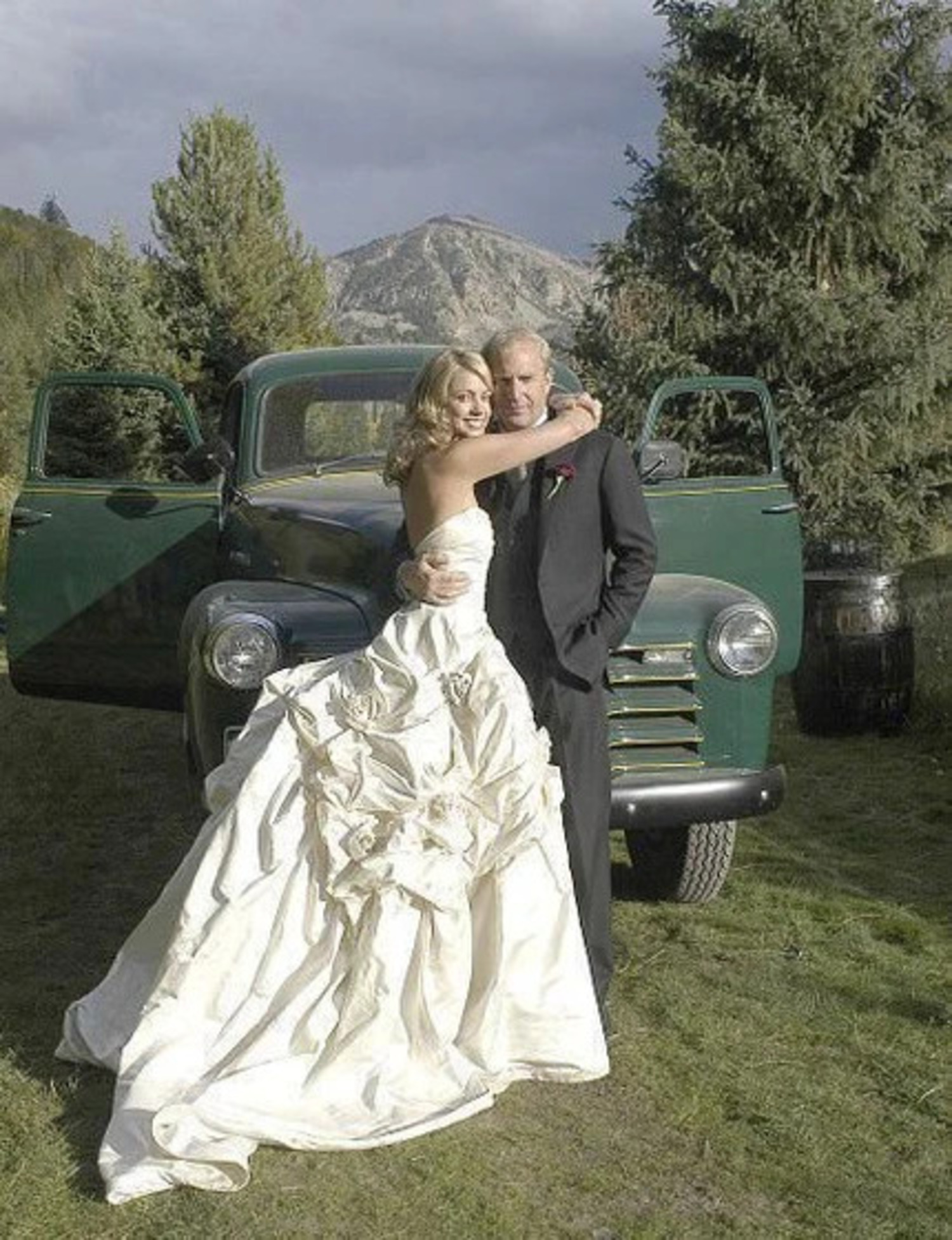 Christine Baumgartner in a ruffled white wedding dress and Kevin Costner in a black three-piece suit with a green vintage car and a mountain as their background on their wedding