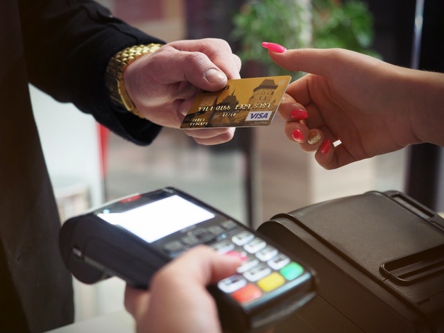 Person wearing gold watch hands Visa credit card to cashier wearing pink manicure and holding a point-of-sale terminal