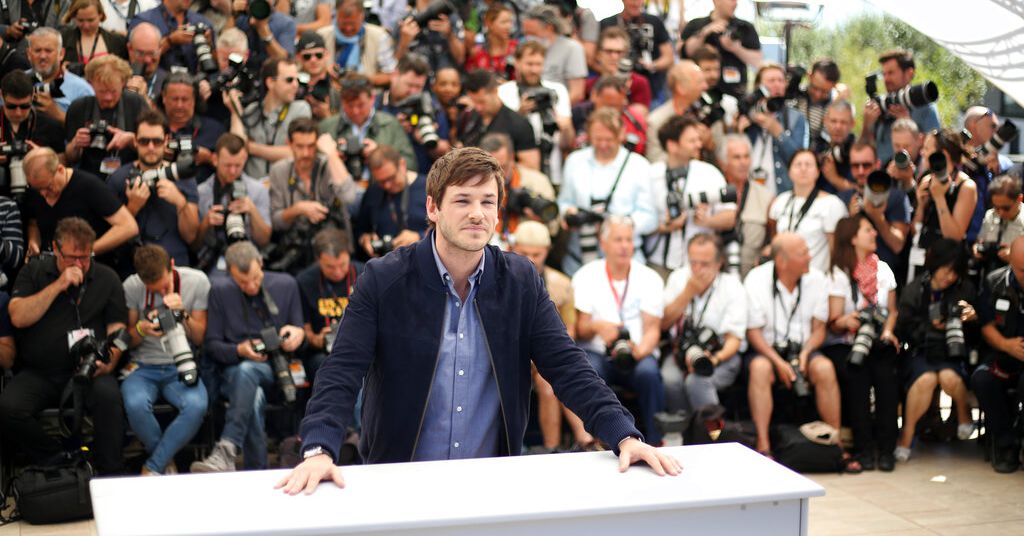Gaspard ulliel is standing in a blue jacket with hands on a table while crowd of photographers sit in the back