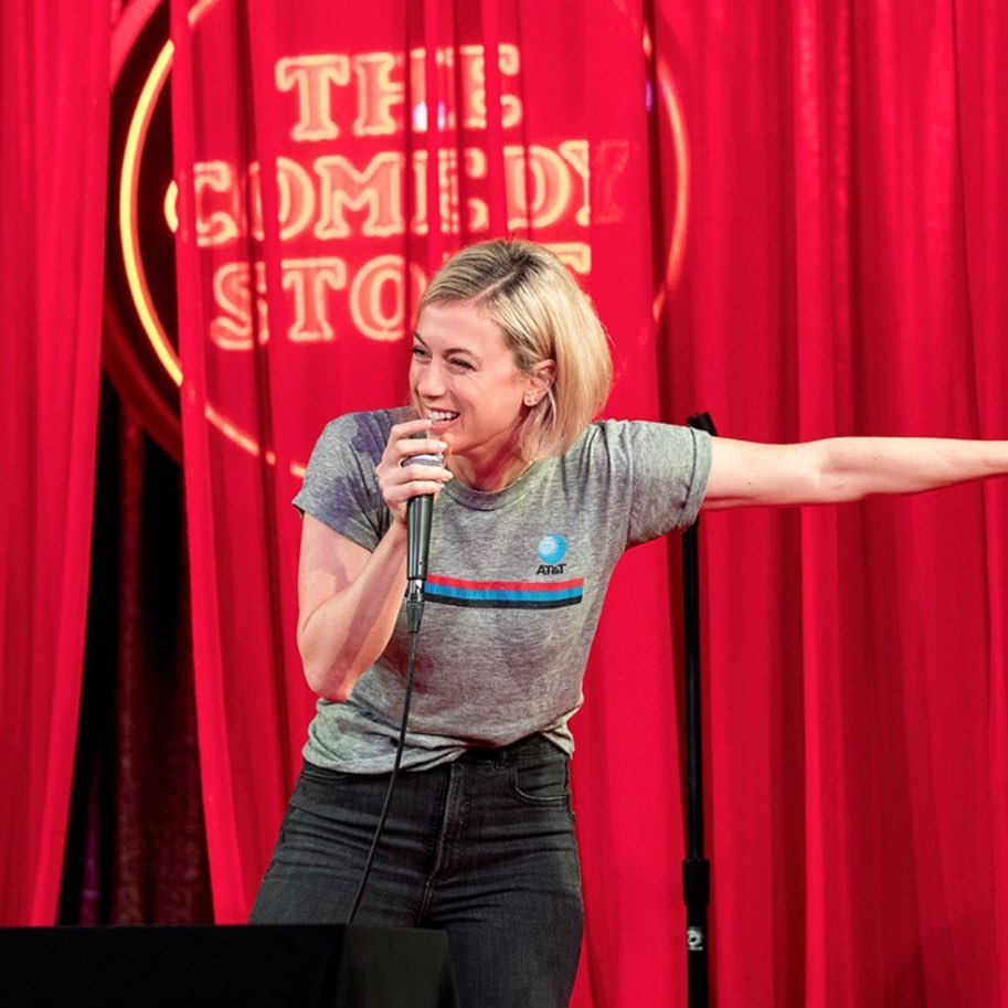 Iliza Shlesinger in an AT&T shirt performing on stage, with a red curtain and a sign saying ‘The Comedy Store’ as backdrop