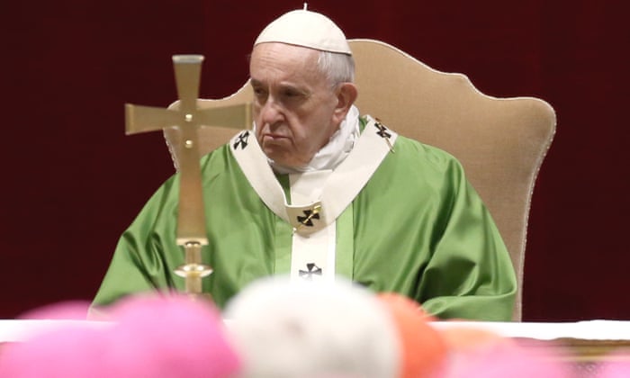 Pope benedict wearing a green and white-colored big gown sitting on a chair in front of a cross sign