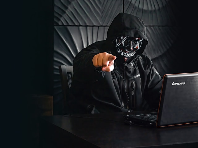 Masked hacker using Lenovo laptop in dark room points his finger at the camera
