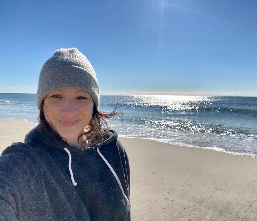 Elisa Pugliese in a long sleeve hooded sweatshirt and ribbed wool beanie takes a selfie at the beach in New York