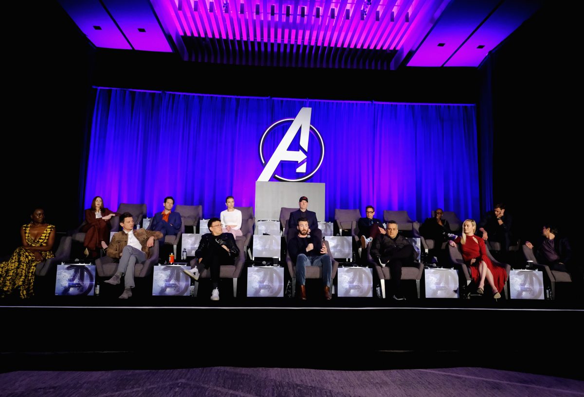 The Avengers performers are seated on the stage one seat apart, with the Avengers logo in the center of the violet curtain 