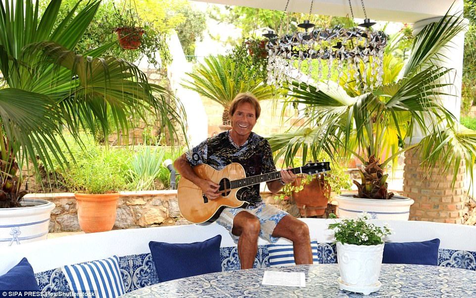 Sir Cliff Richard looks very happy while playing a guitar on a patio with a lot of plants