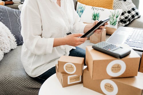 Parcels, calculator, and a laptop in front of a woman holding her tablet while tracking her online business