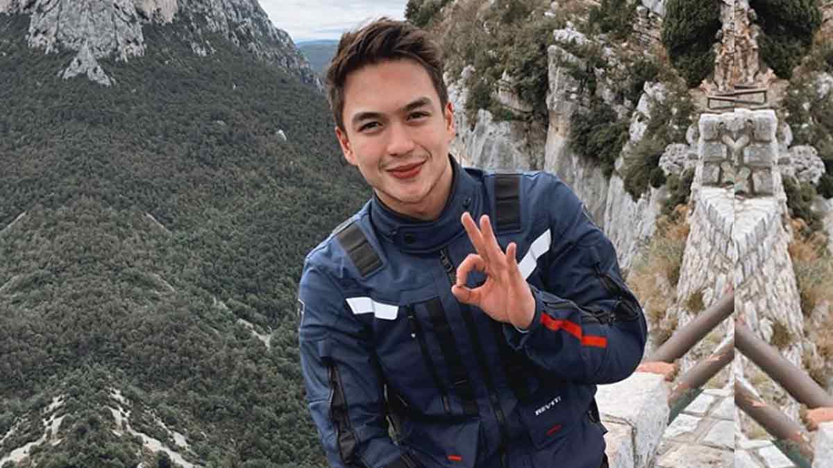 Dominic roque wearing a blue jacket while doing a satisfactory sign with his right hand on top of a mountain