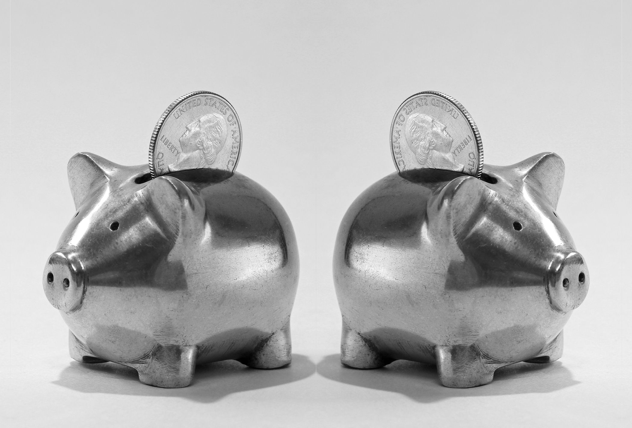 Two silver piggybanks with one silver coin above it on the coin slot