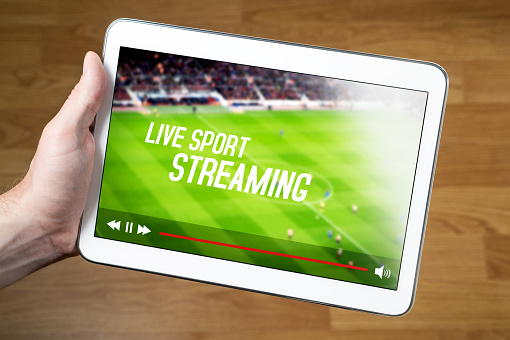 A person holding a white tablet with a live sports streaming video on it