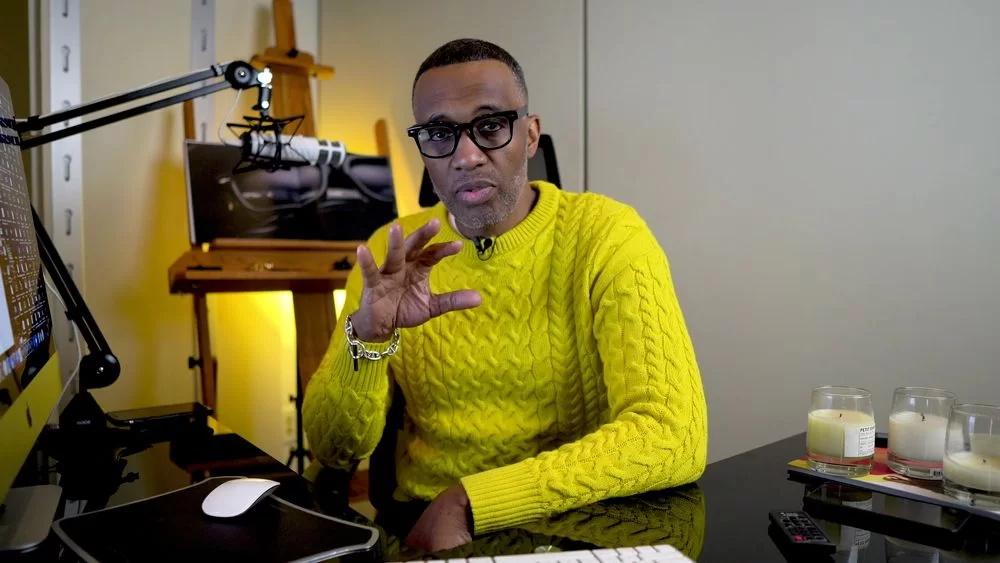 Kevin Samuels in yellow top and black glasses looks while sitting in his office