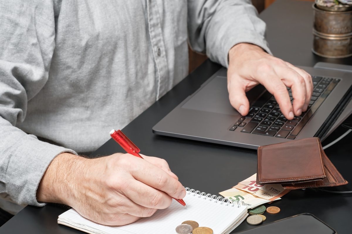 A man writing in a notebook and a laptop, wallet, money, and coins in front of him