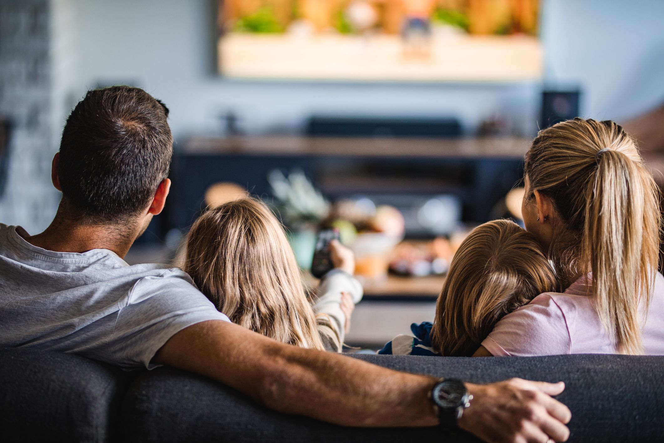 A family is sitting on the couch, and the little girl in the middle is holding the remote