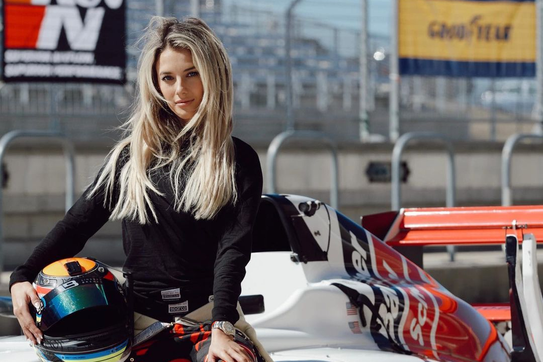 Lindsay Brewer sitting in a race car while wearing black long sleeves and silver watch and holding a helmet