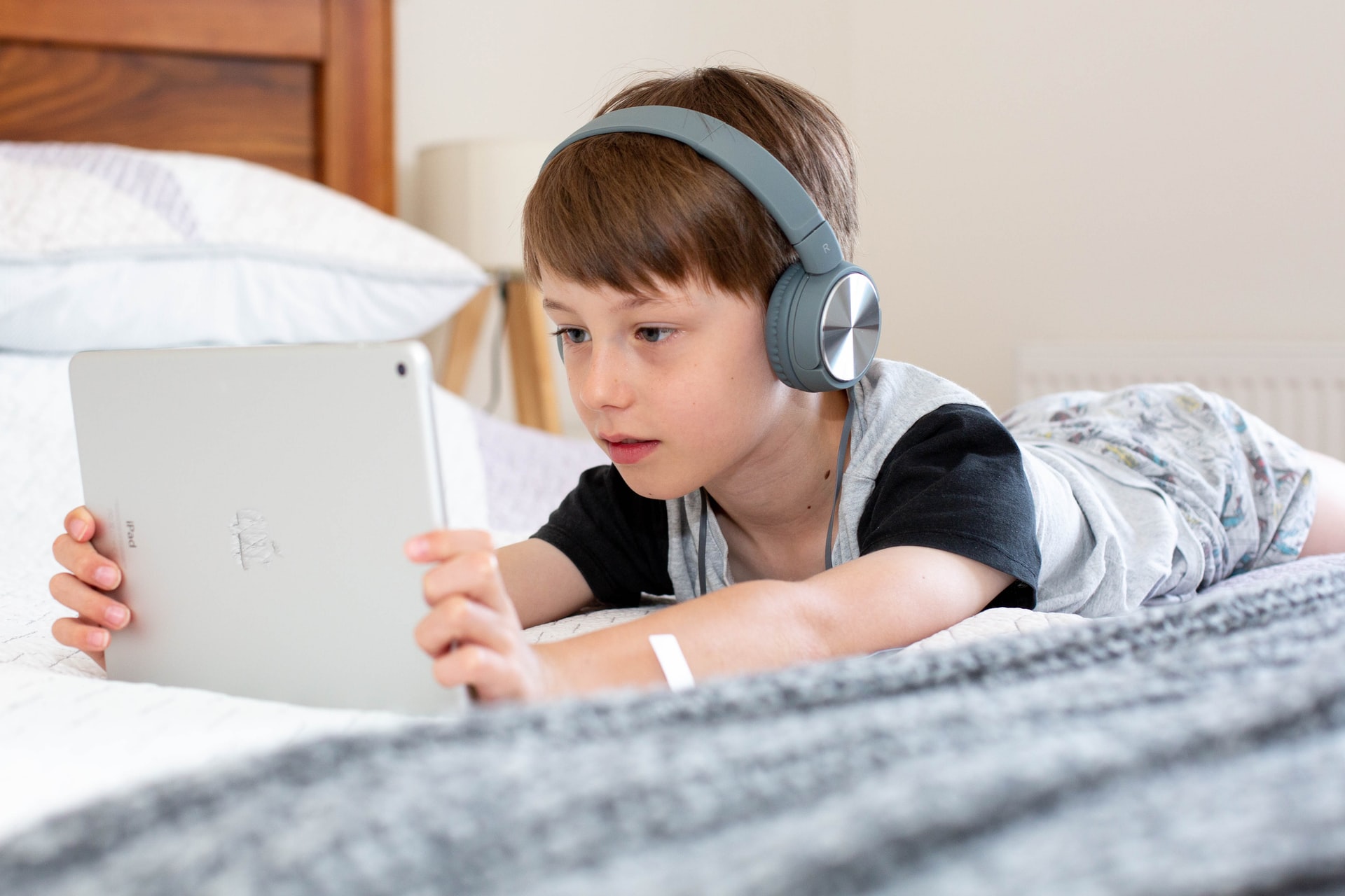 A boy lying in bed on his belly watching cartoons online on his iPad with headphones on