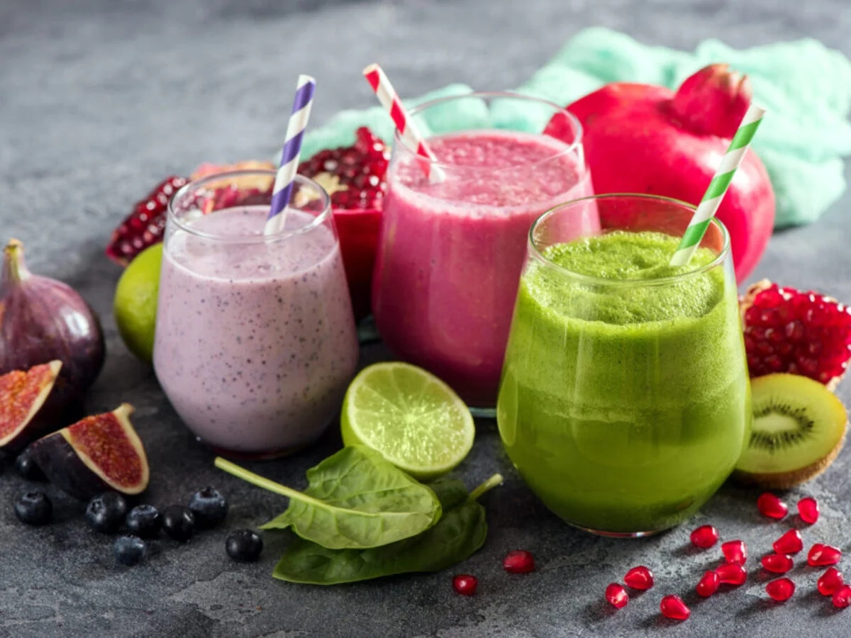 Green smoothie, purple smoothie and pink smoothie with some fruits and vegetables placed on a grey surface