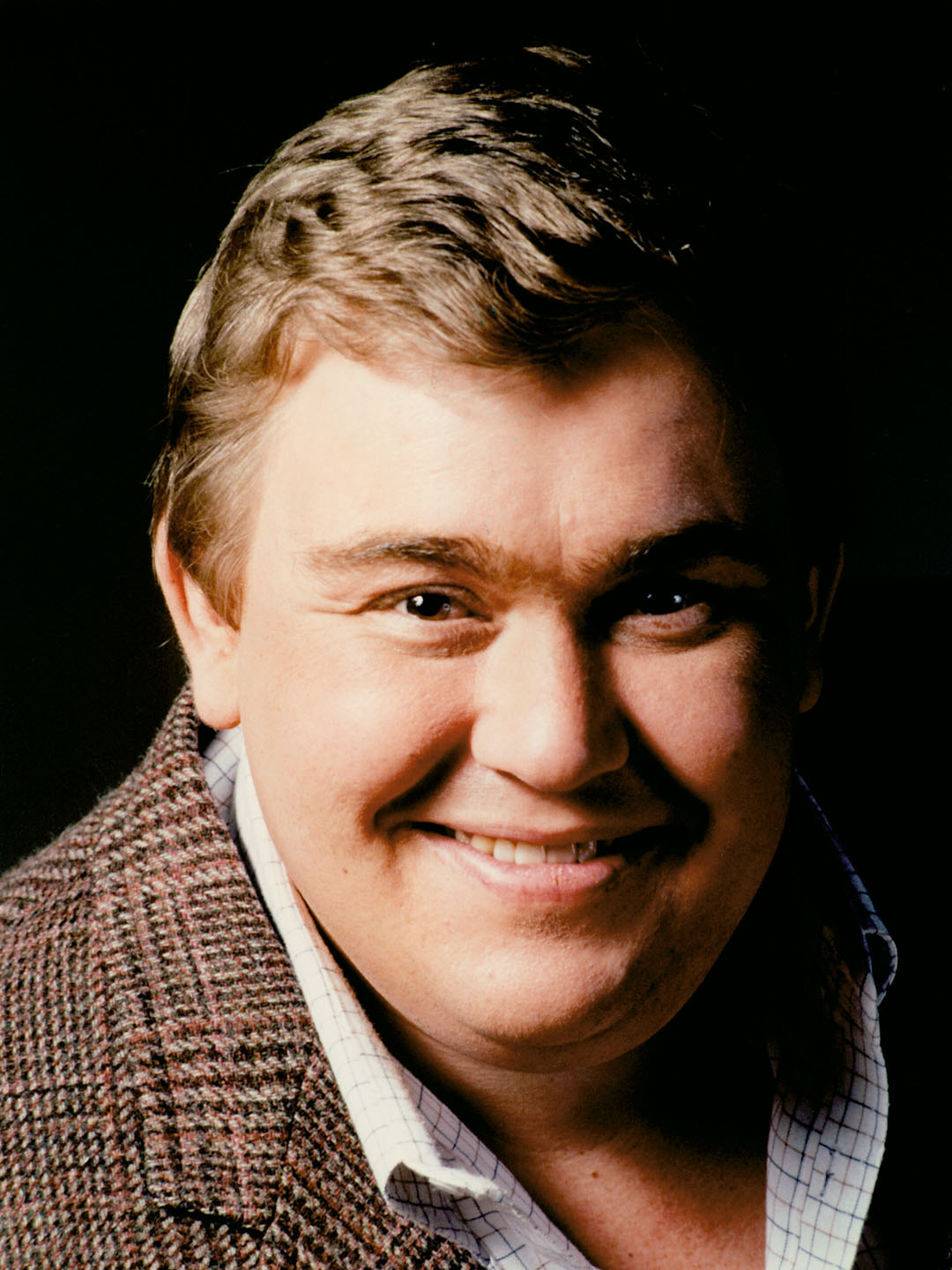 John Candy wearing a brown coat and smiling
