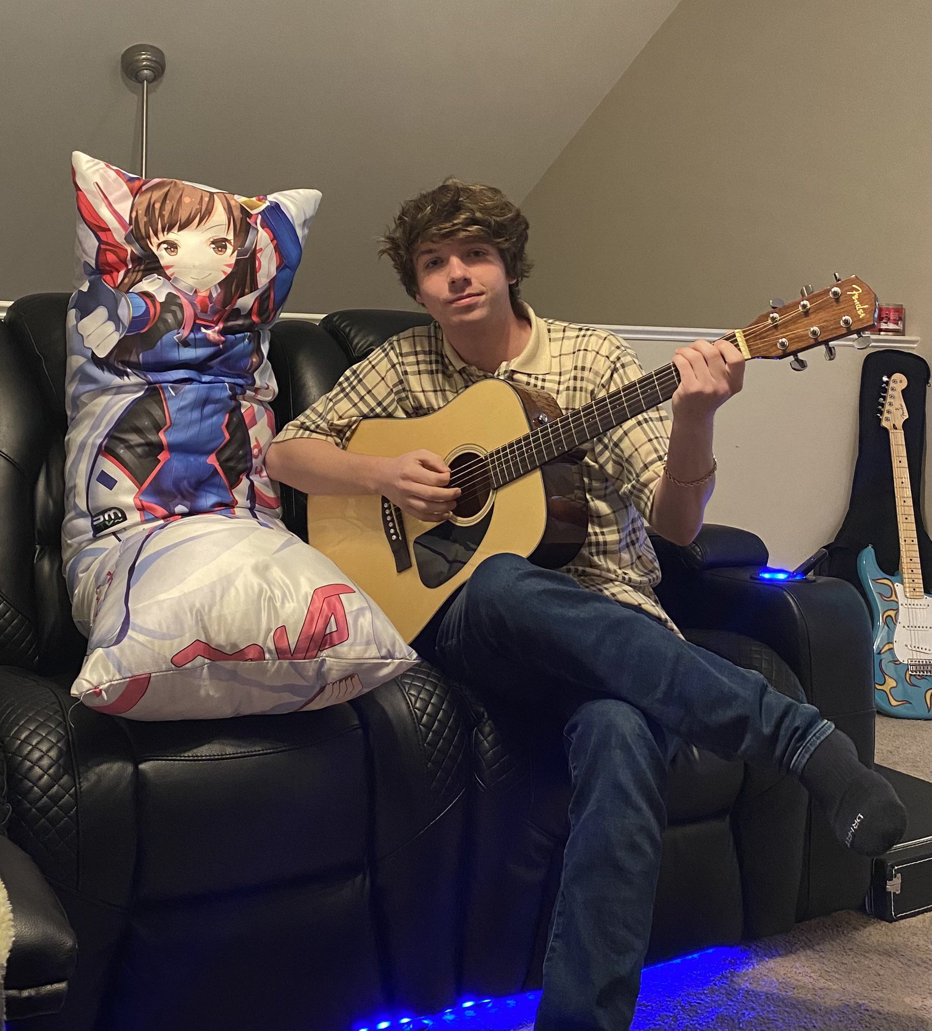 Karl Jacobs holding a guitar and wearing checkered shirt with an anime girl pillow 
