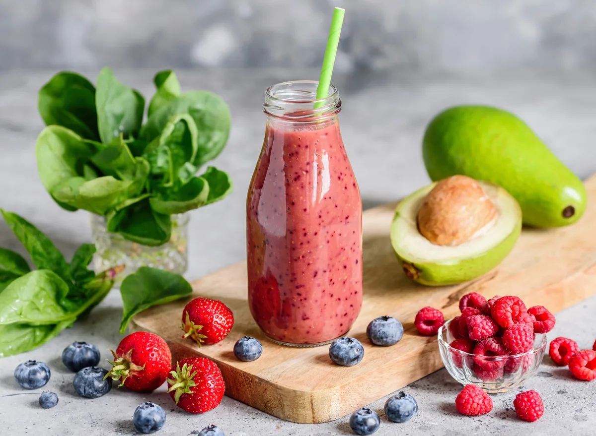 Red smoothie in a glass bottle with avocado, berries, and spinach on a grey surface
