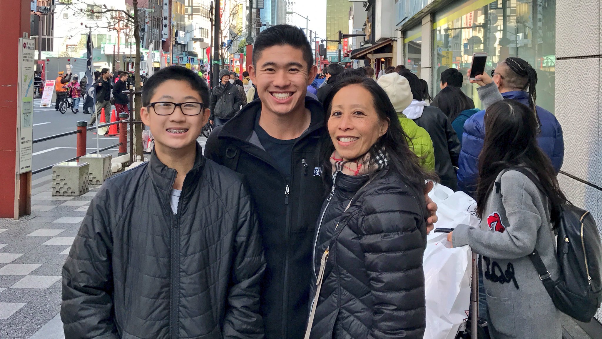Debbie Morikawa wearing a black jacket and checkered scarf with her sons, Collin and Garrett smiling