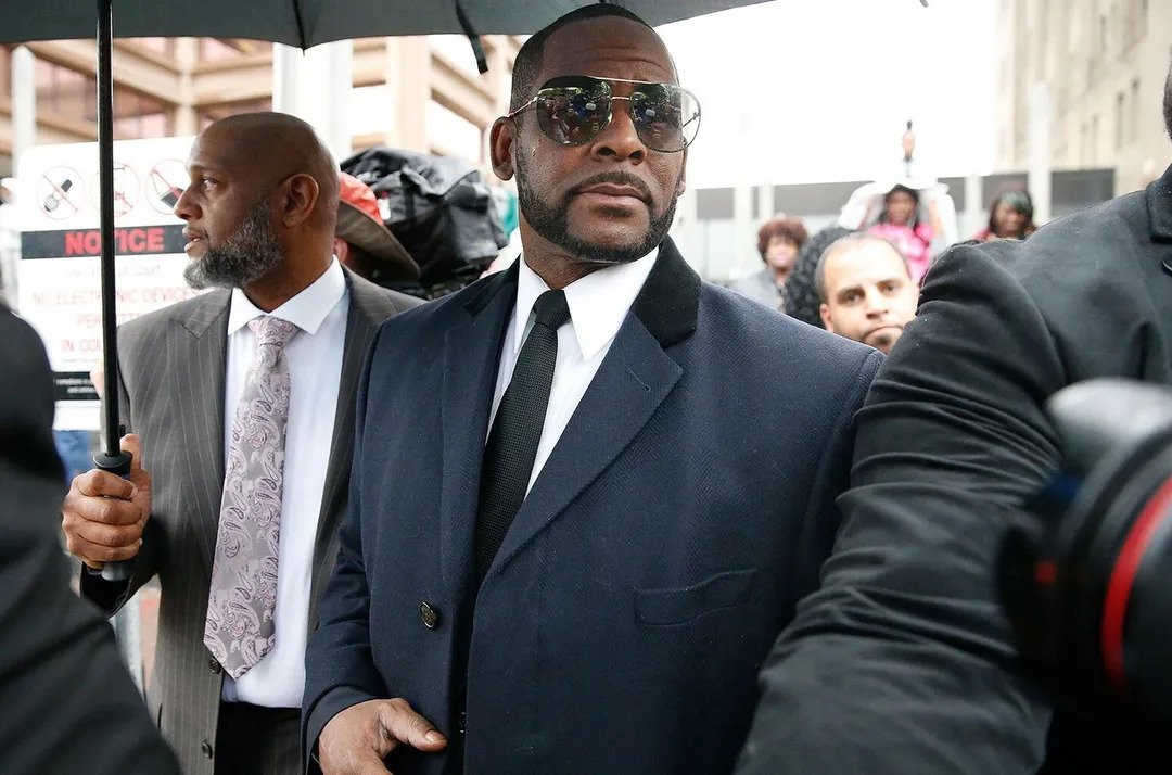 A bald man holds an umbrella for R. Kelly who is in a navy suit and dark glasses
