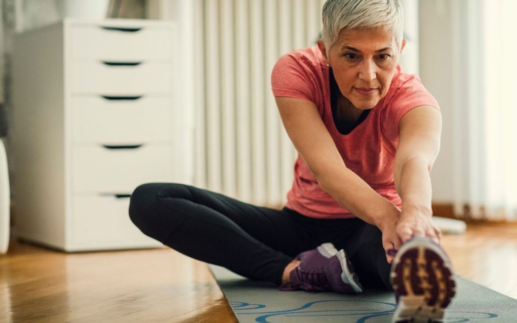A gray-haired woman is doing some stretching in her room