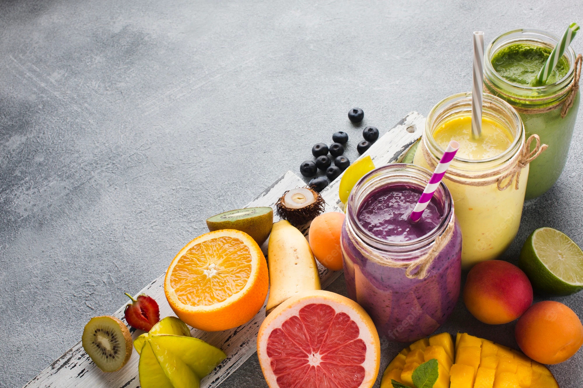 Purple smoothie, yellow smoothie and green smoothie in glass jars with fruits placed on a grey surface