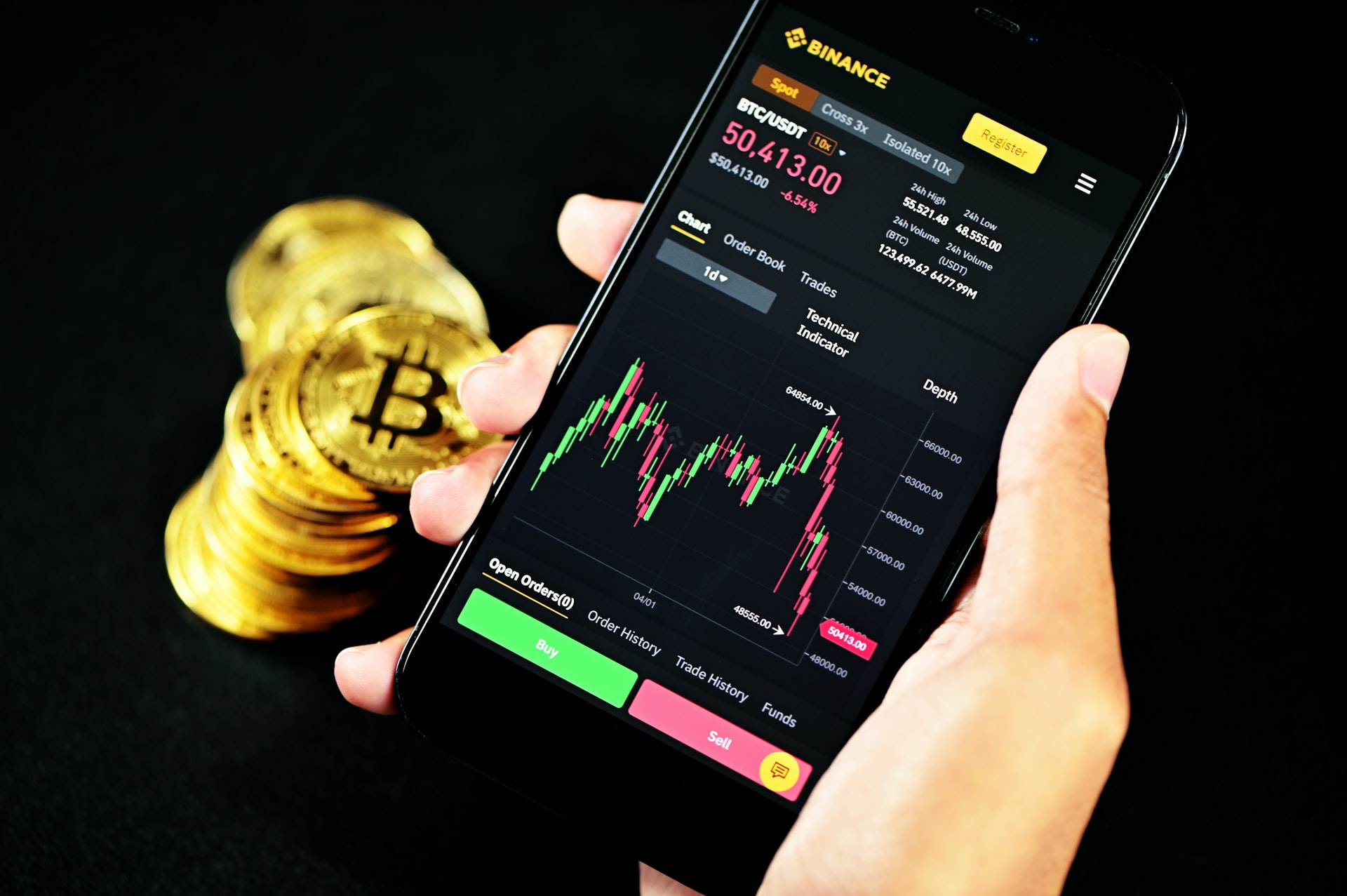 A stack of Bitcoins and a smartphone held by a hand showing Bitcoin trading on the Binance website 