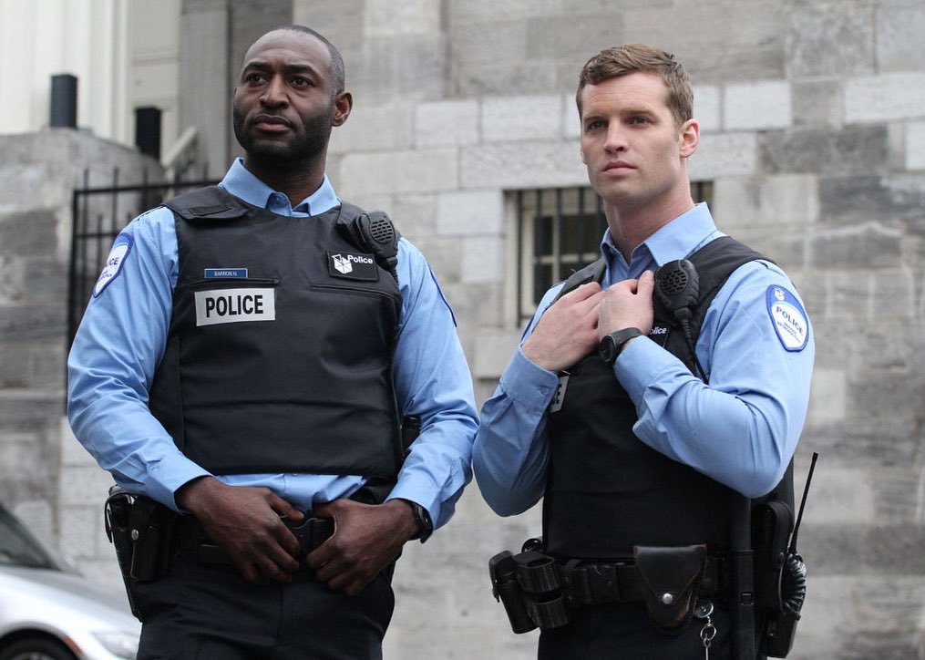 Adrian Holmes and Jared Keeso in light blue police uniform with black vest
