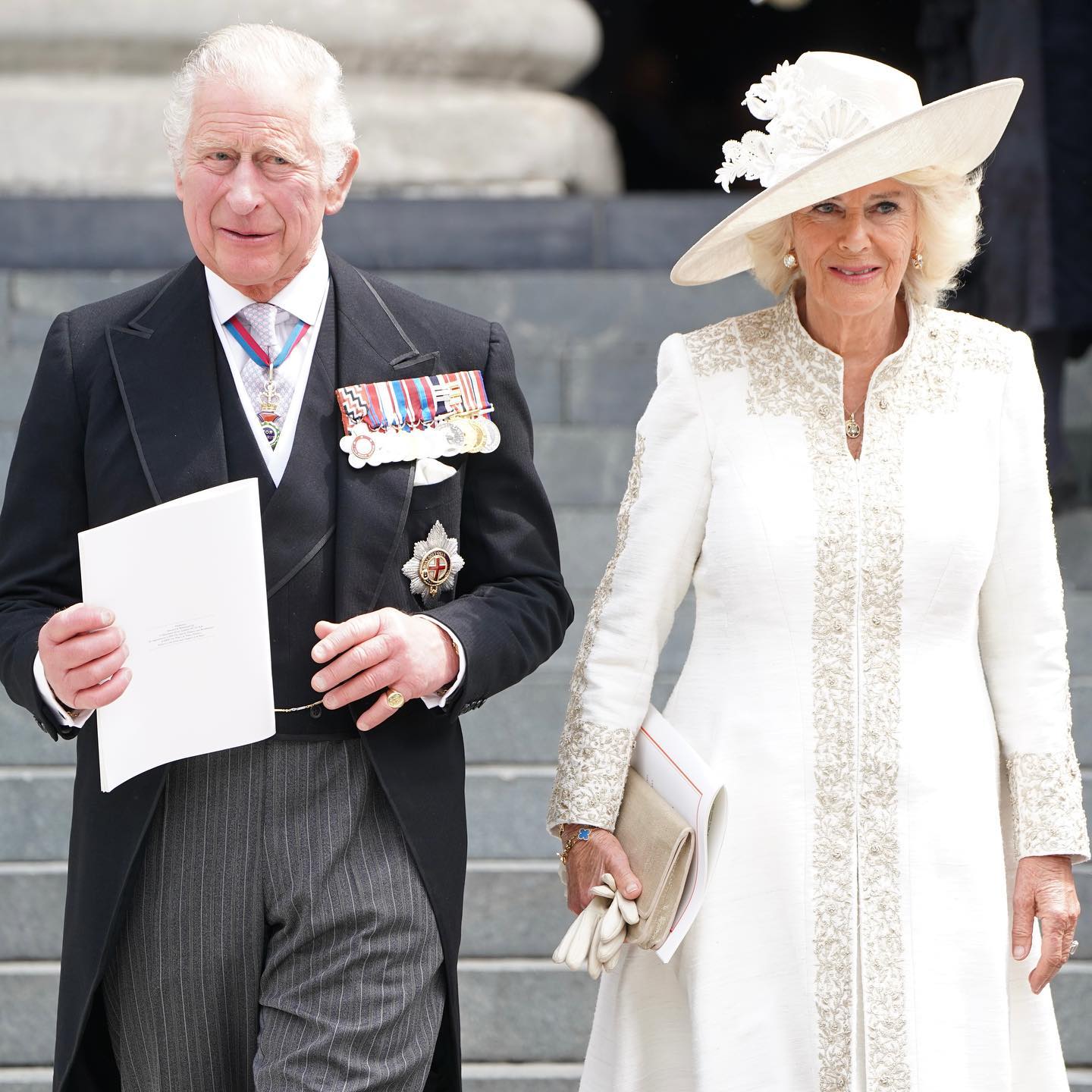 King Charles III in black tailcoat with pinned medals and Camilla in white wide brimmed hat and clothes