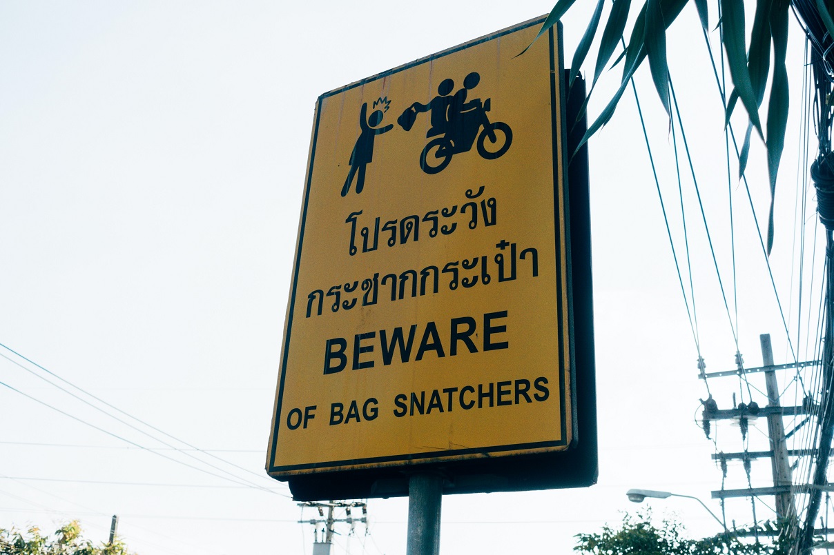A vertical sign in pale yellow metal plate with the words, beware of bag snatchers