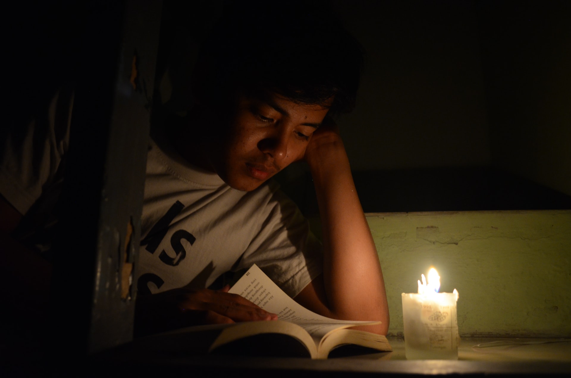 A young Asian-looking man with head resting on an arm reads a book in front of a burning wide white candle