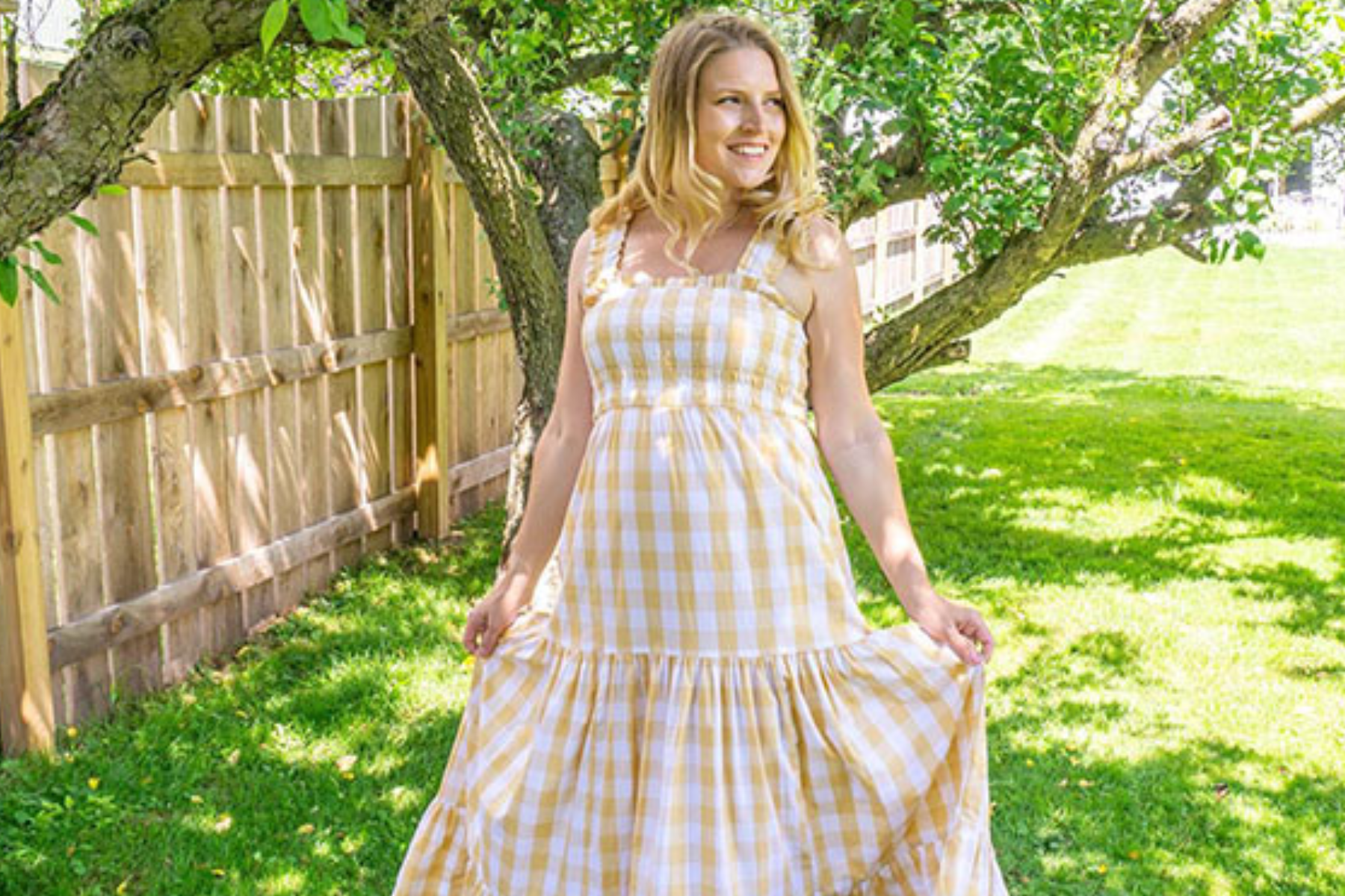 A woman in a yellow checkered dress poses in her backyard