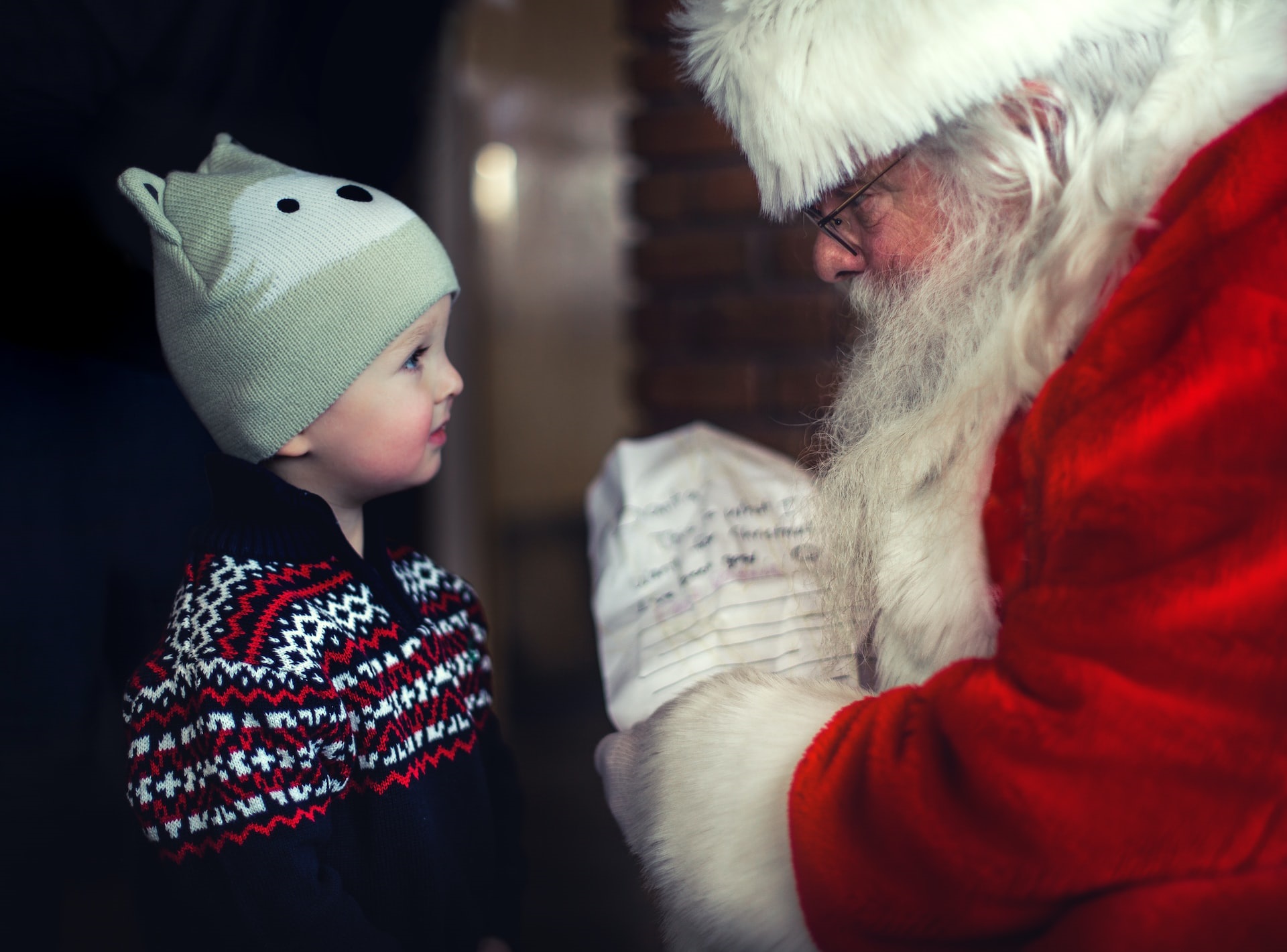 A male toddler in black sweater and beanie looks at Santa Claus holding a paper