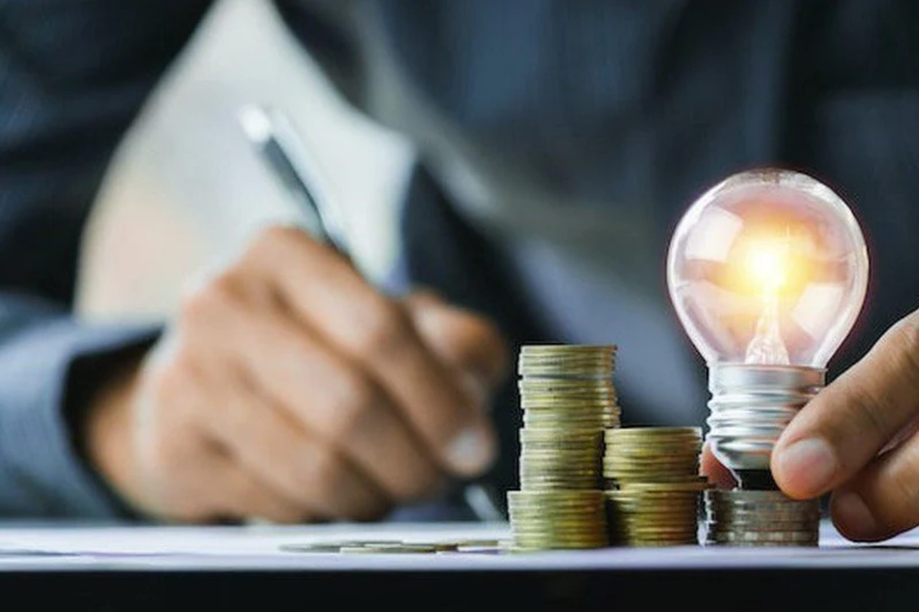 A man writes on a piece of paper while holding a little light bulb and some coins