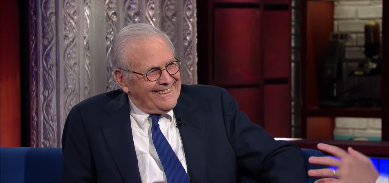 A smiling Donald Rumsfeld in white shirt under a navy blue blazer and blue tie at The Late Show with Stephen Colbert