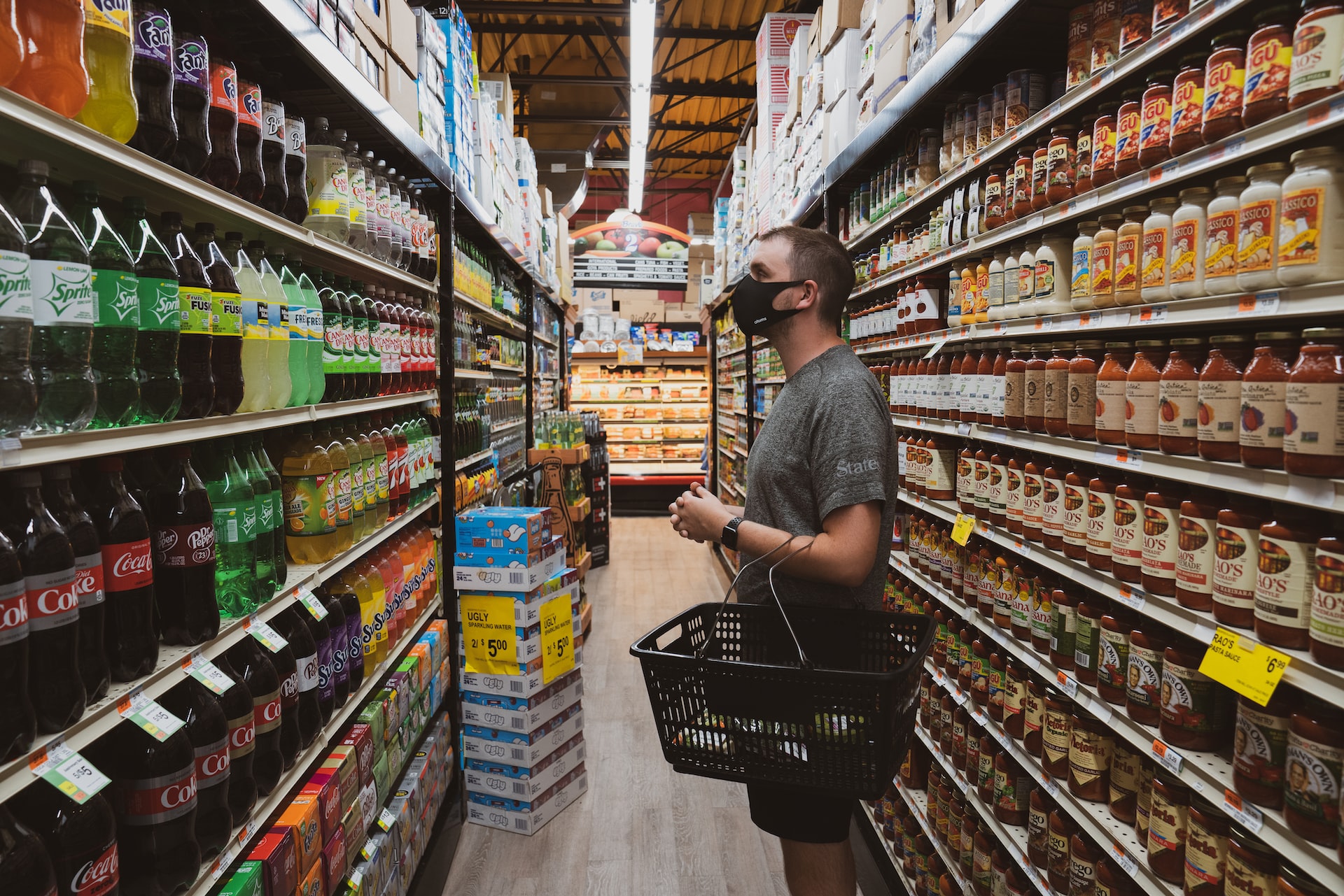 A man in gray T-shirt and black face mask carrying a black grocery basket and checking out beverages