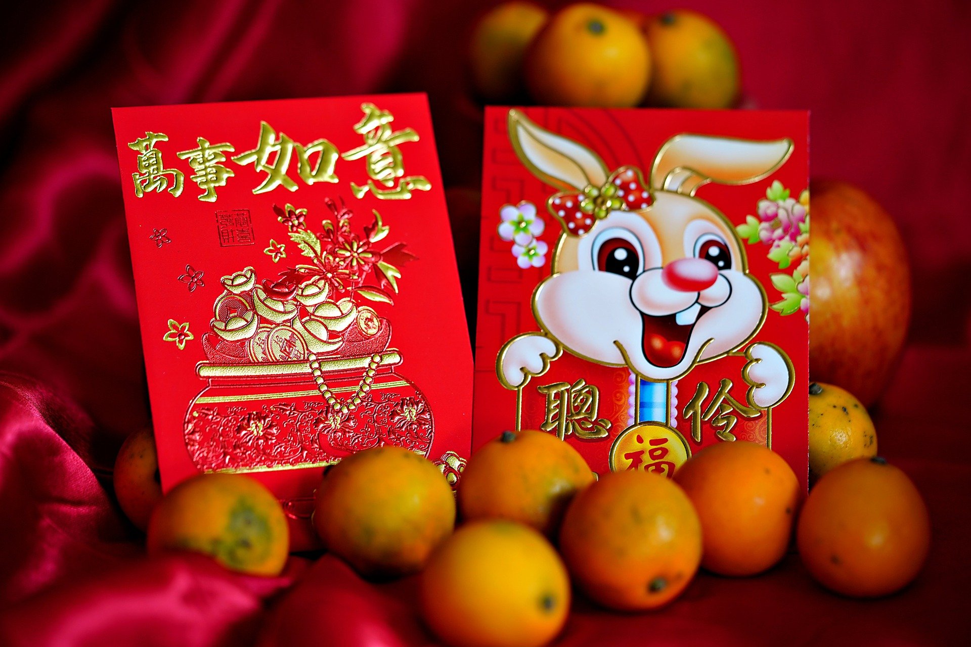 Two red packets with one showing a smiling rabbit standing among an apple and some kumquats