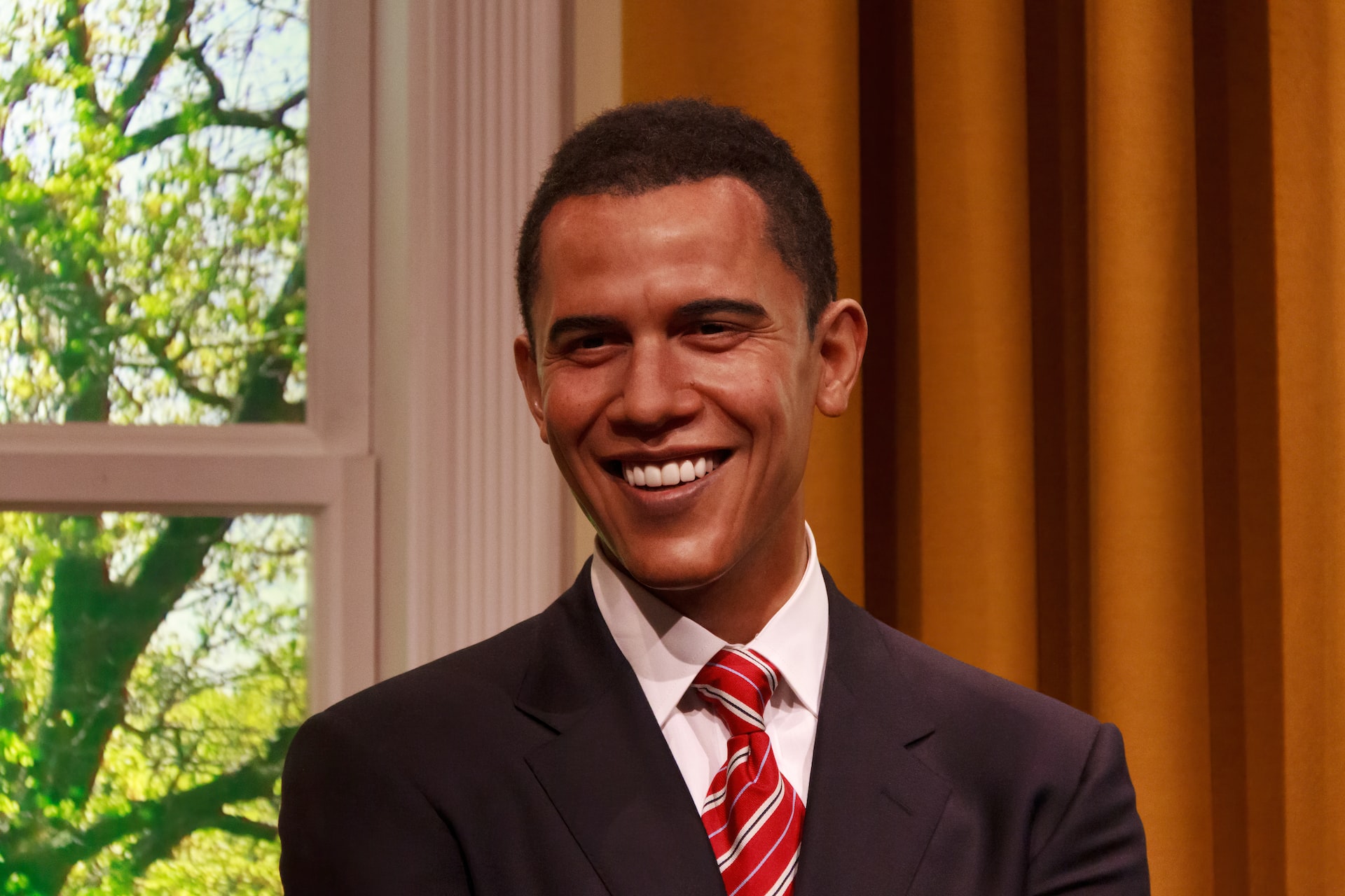 Wax figure of a smiling Barack Obama in dark suit and red tie standing by a window